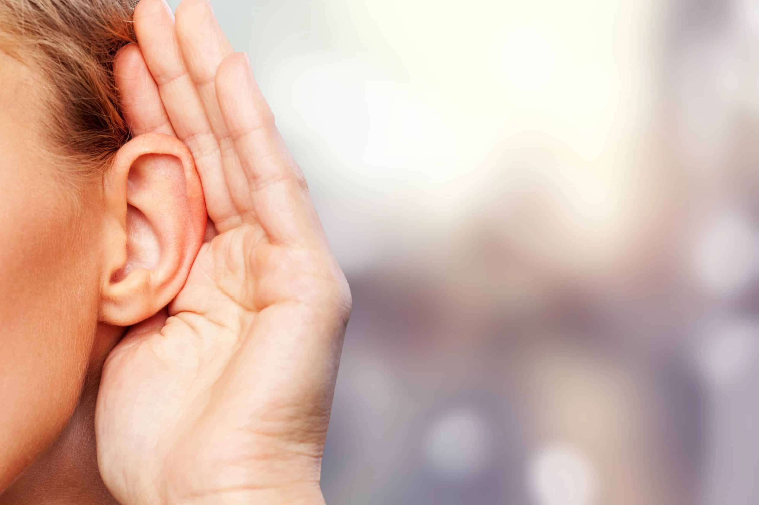 A woman cupping her ear with her hand, leaning slightly forward with a curious expression, symbolizing difficulty hearing or early signs of hearing loss.