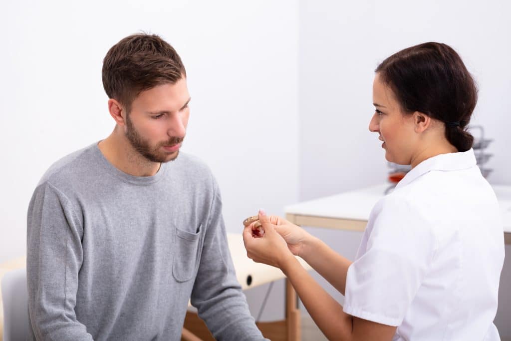 Young Male Patient Looking At Female Doctor Holding Hearing Aid In Clinic